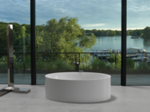 Freistehende, ovale Badewanne vor Panoramafenster mit Blick auf See, Steg und grüne Uferlandschaft.
